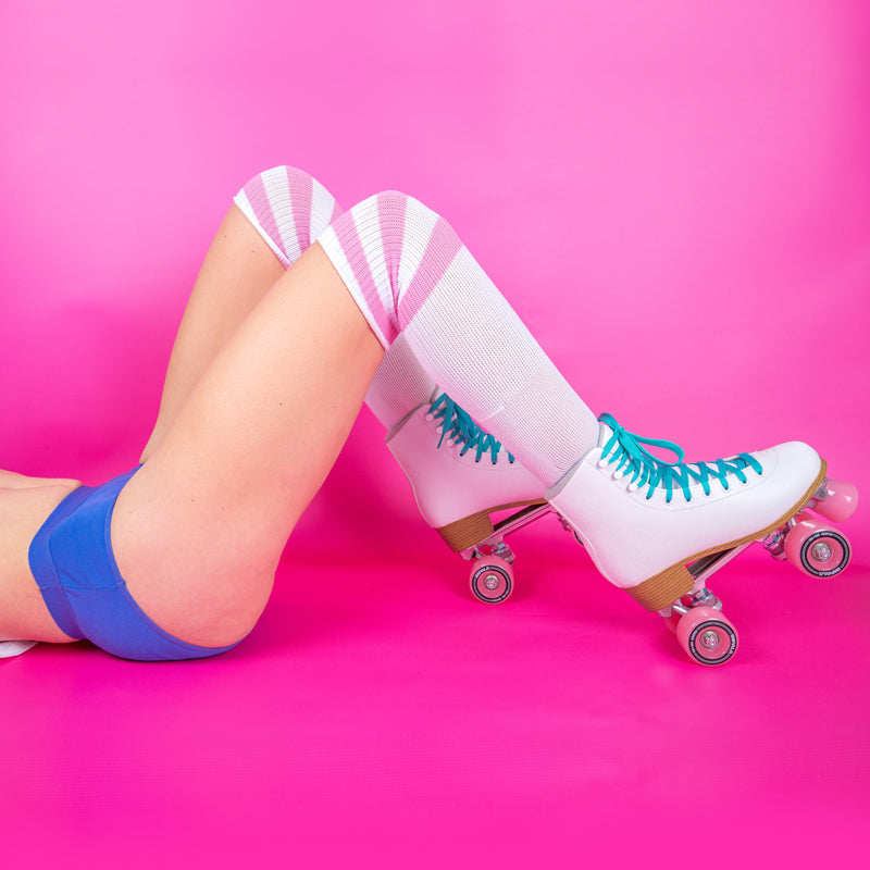Model wears blue hipster period pants.   The background is hot pink.  Model is laying in her back.  She also wears long white and pink socks and white roller boots with pink wheels and turquoise laces.  Her legs are bent at the knee and torso not visible.