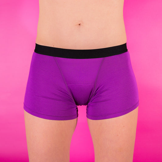 Model wears purple period pant boyshorts with a black waistband.  The model faces front and the background is hot pink.