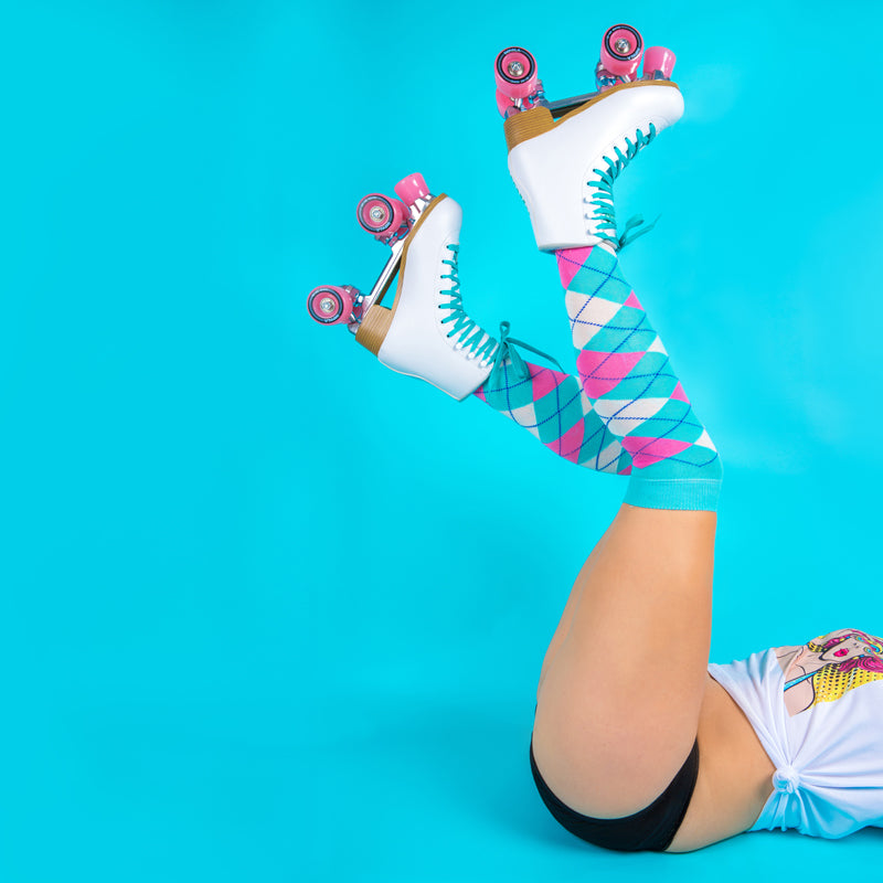 She wears back period pants with a lace waist band and a white t shirt knotted at the side.  The background is bright turquoise background .  The model is laying on her back with her legs in the air.  She is wearing white roller boots with pink wheels and turquoise laces, long turquoise, pink and white socks.